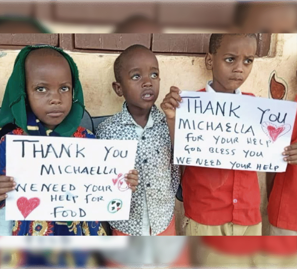 Three children holding handwritten signs thanking Michaella for her help and requesting food assistance, expressing gratitude and need.