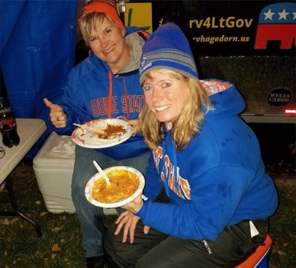 Michaella Franklin in a blue Boise State hoodie and beanie, smiling and holding a plate of food, seated outdoors with another woman also enjoying a meal and giving a thumbs-up.